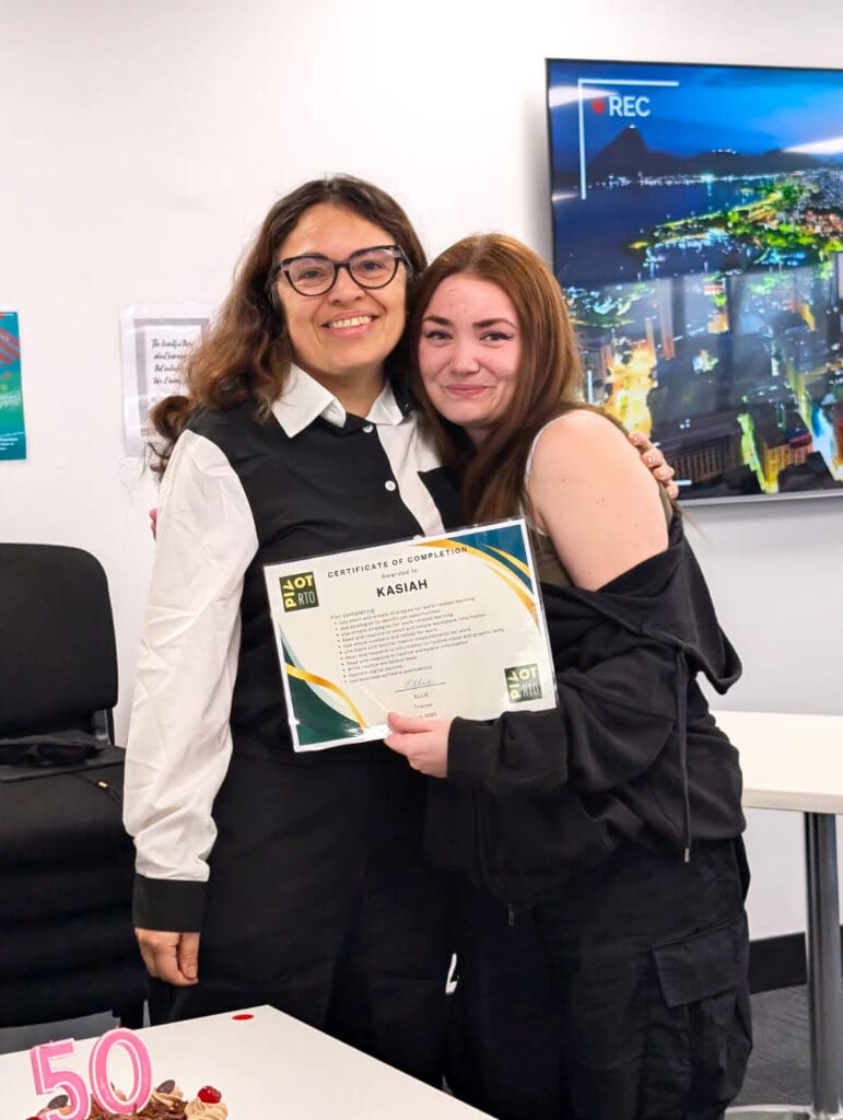 Kasiah smiles as she hugs her trainer Ellie while holding her Certificate I in Skills for Vocational Pathways, marking her achievement in the Skills for Education and Employment Program (SEE program) and her progress in learning for employment whilst exploring Australian career pathways.